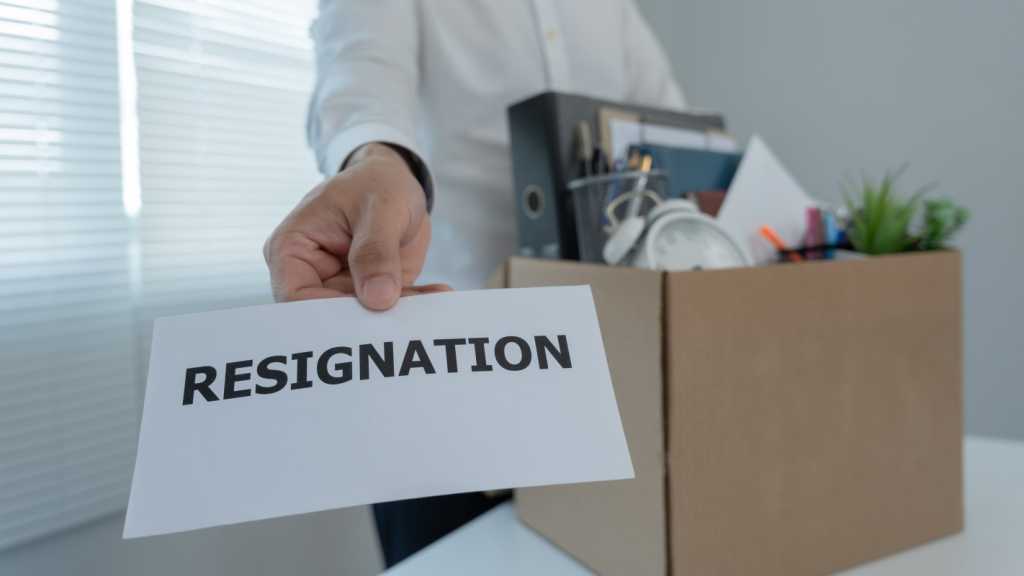 A person holding a piece of paper that says Resignation on it. They are standing next to a desk, which has an assortment of office supplies and a pot plant in it.